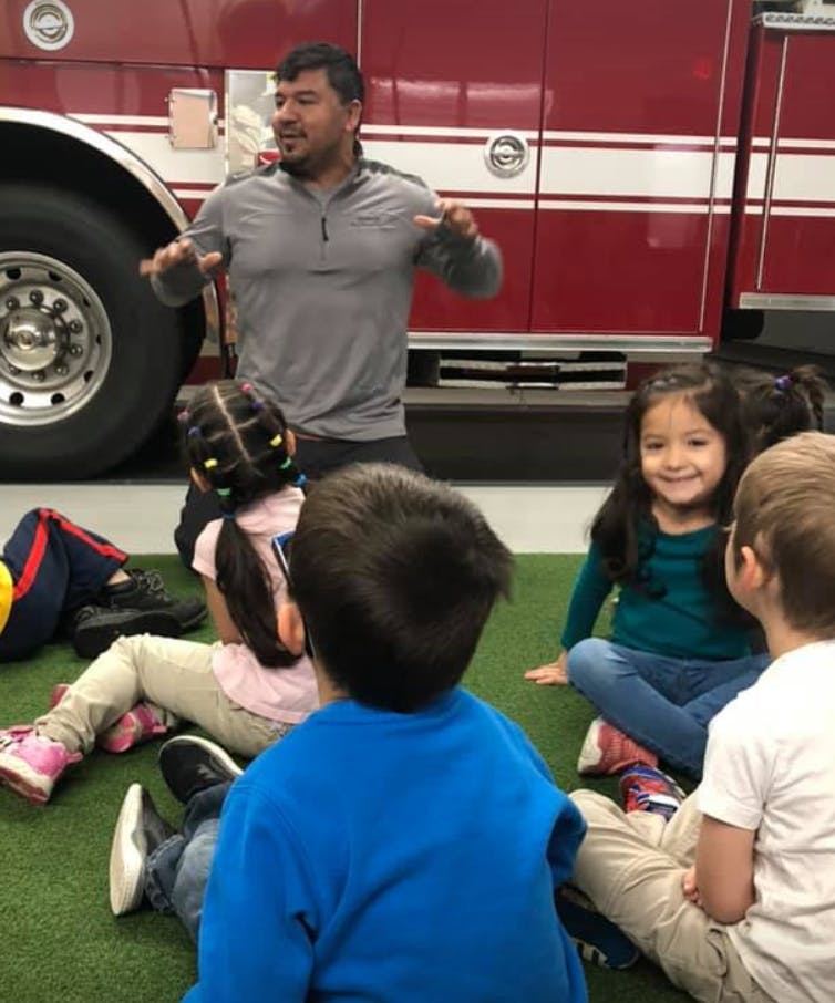 Person speaking to children sitting on the ground in front of a fire truck