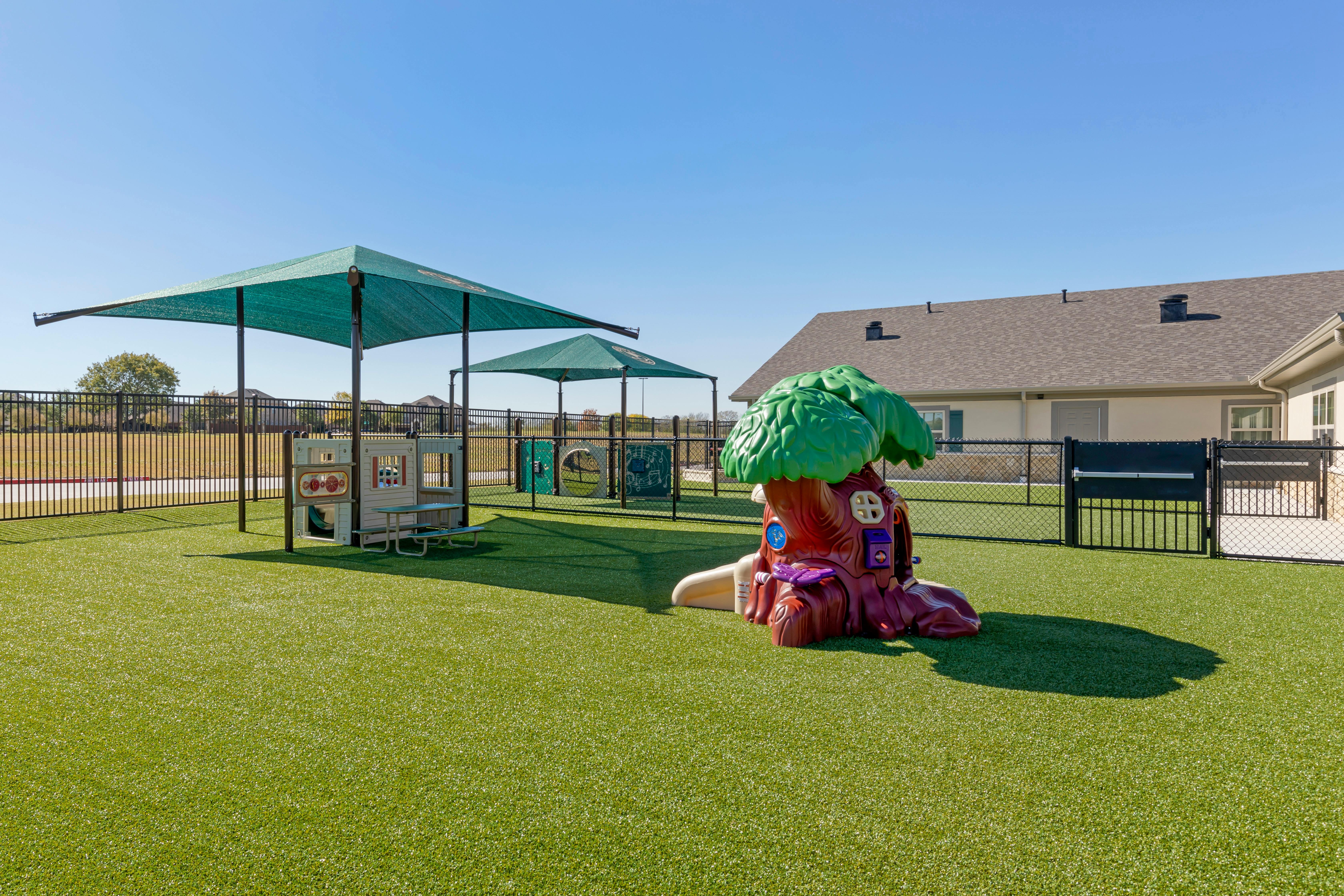 Playground with green turf, a playset, and a tree-shaped climbing toy under shade covers