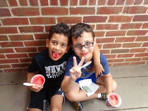 Two children sitting, one holding a cup of ice and the other making a peace sign