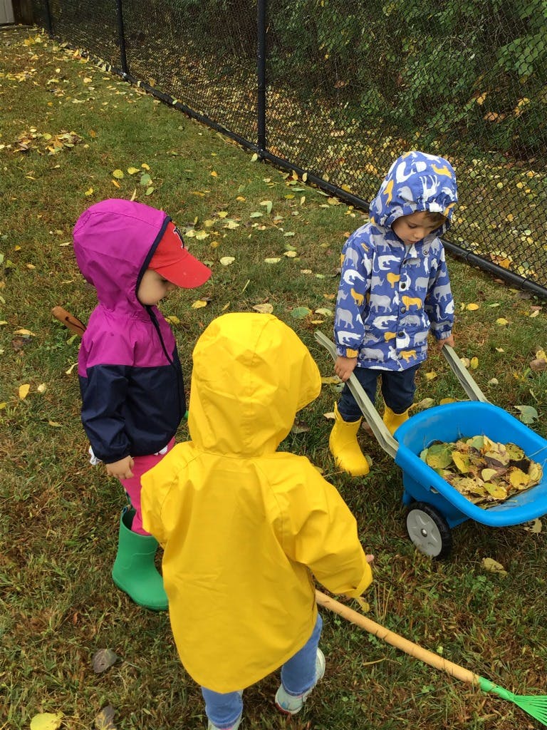 Three children in rain gear raking leaves and pushing a toy wheelbarrow