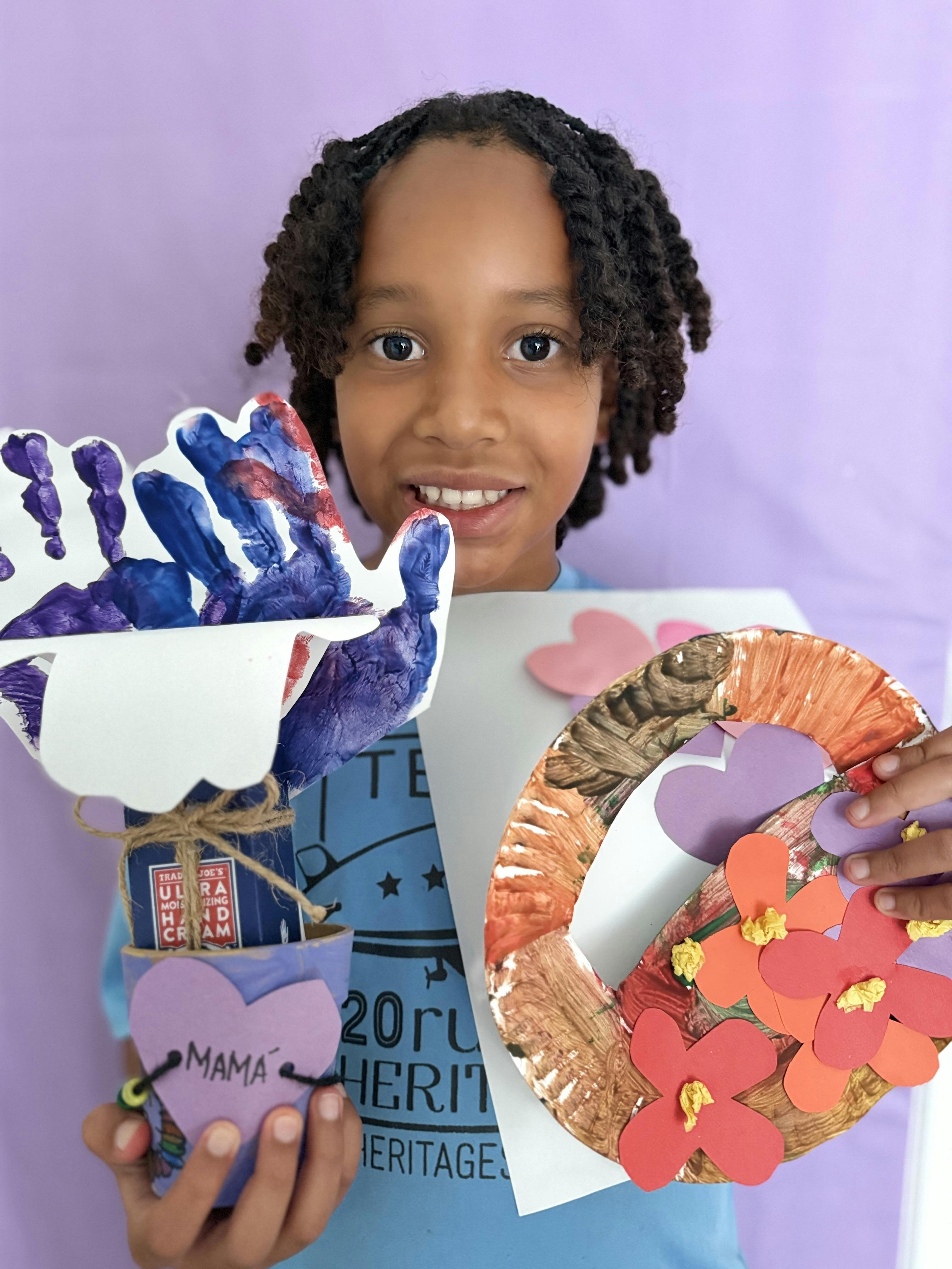 Child holding handmade paper crafts and a small potted gift with a heart tag that says "MAMA"