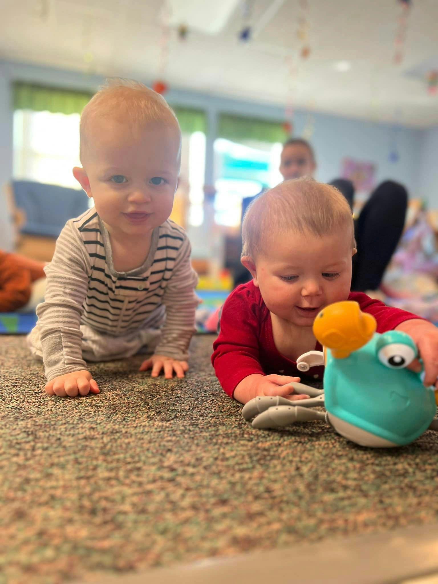 Two babies on a carpeted floor, one crawling and one playing with a toy