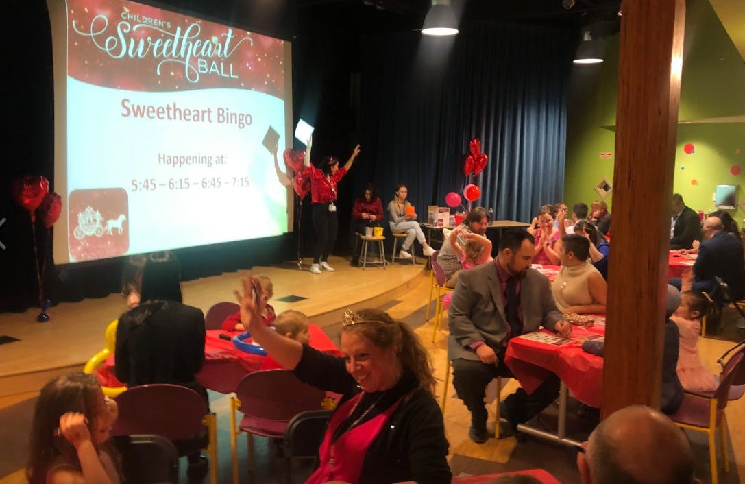 People seated at tables in a room with a large screen displaying "Sweetheart Bingo"