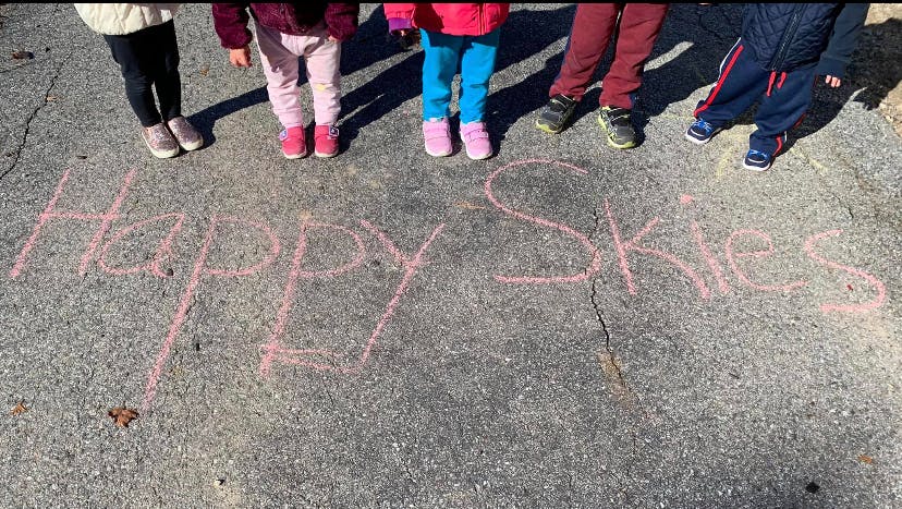 Children standing over chalk writing "Happy Skies" on pavement