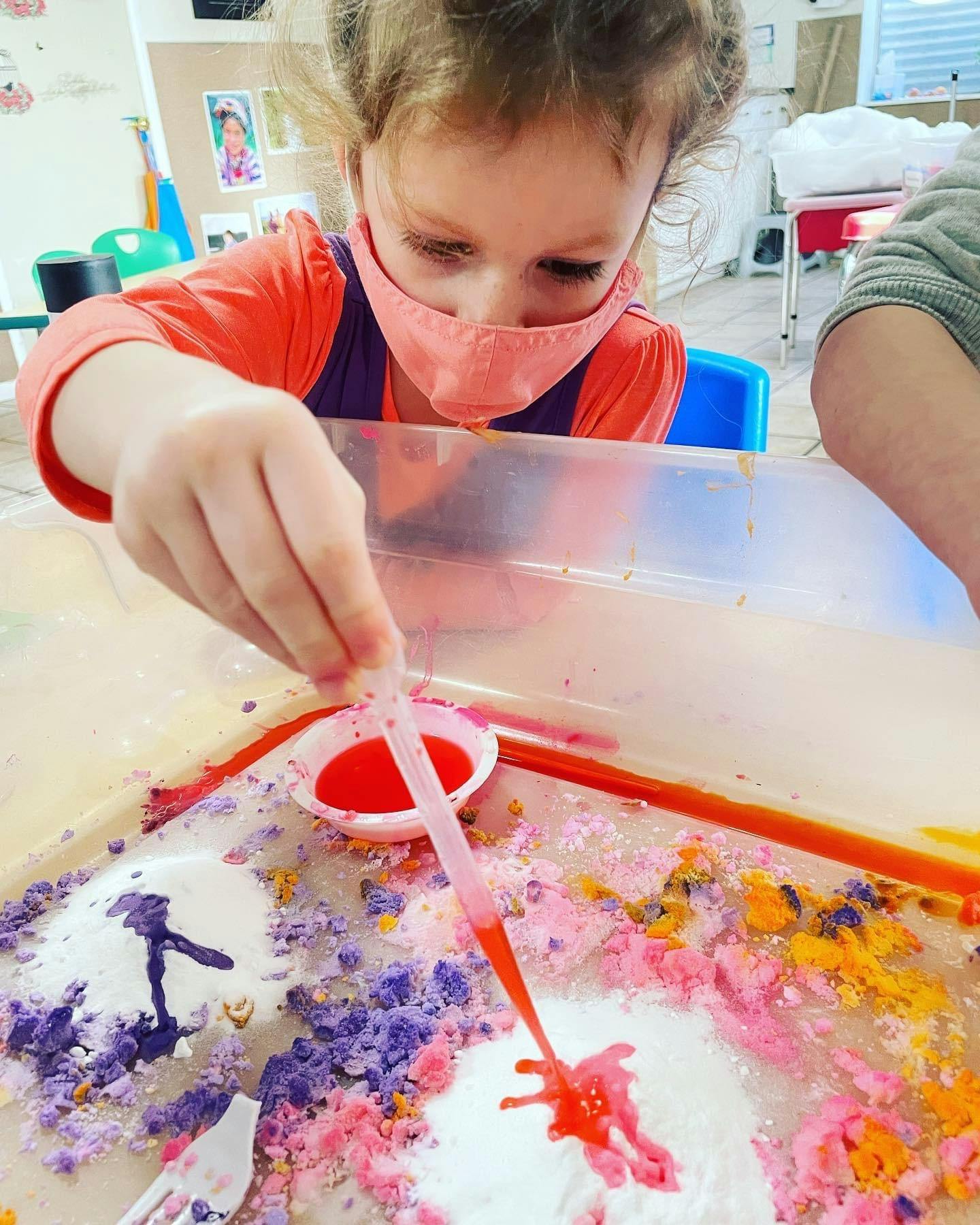 Child in a mask using a dropper with colorful materials in a sensory bin