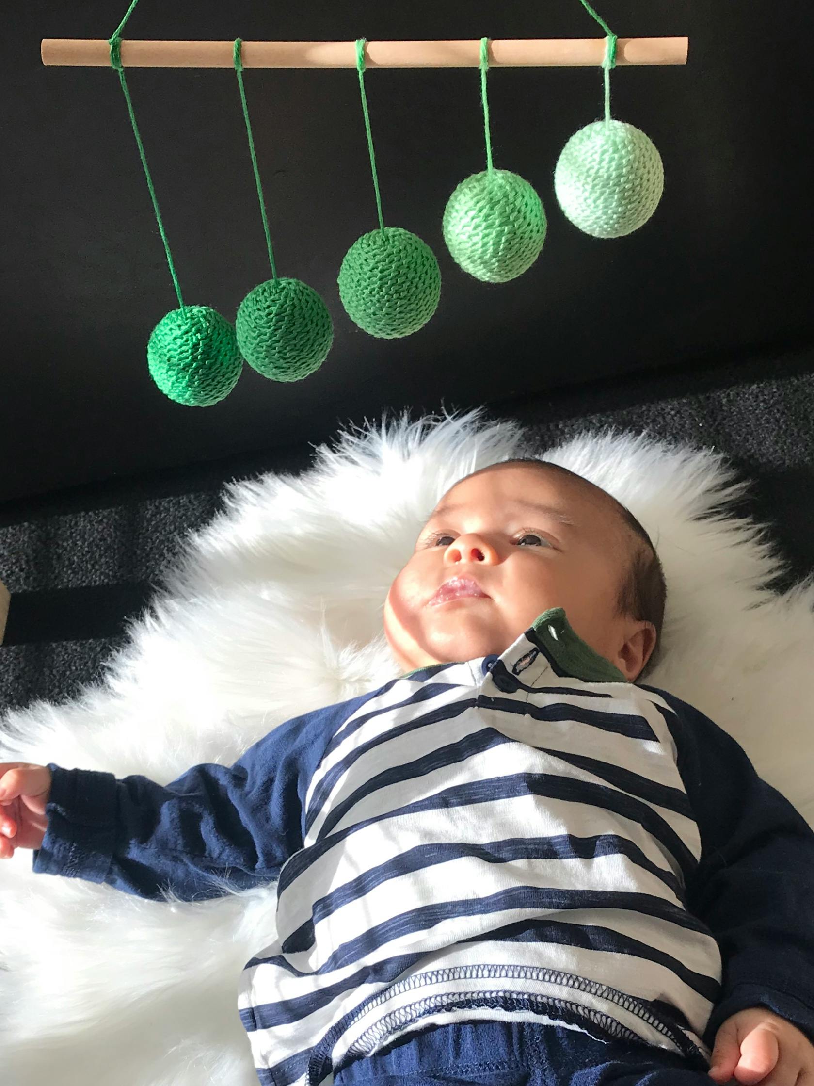 Child lying on a white fluffy blanket looking up at green yarn balls hanging from a stick