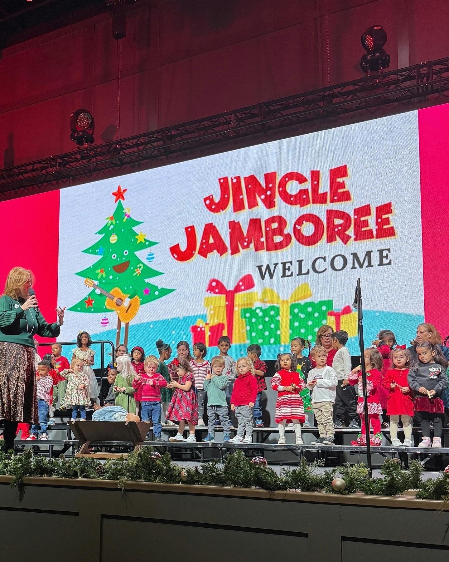 Person speaking to a group of children on a stage with a "Jingle Jamboree" sign