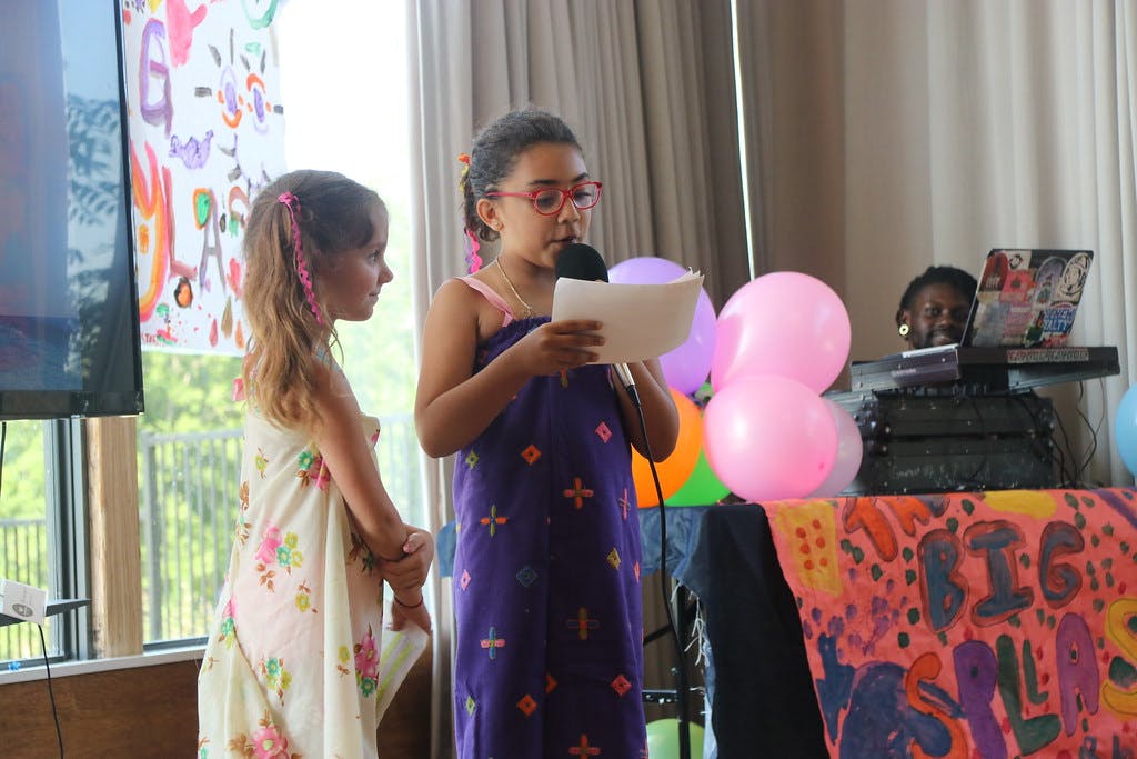 Child speaking into a microphone while holding a paper, another child standing nearby