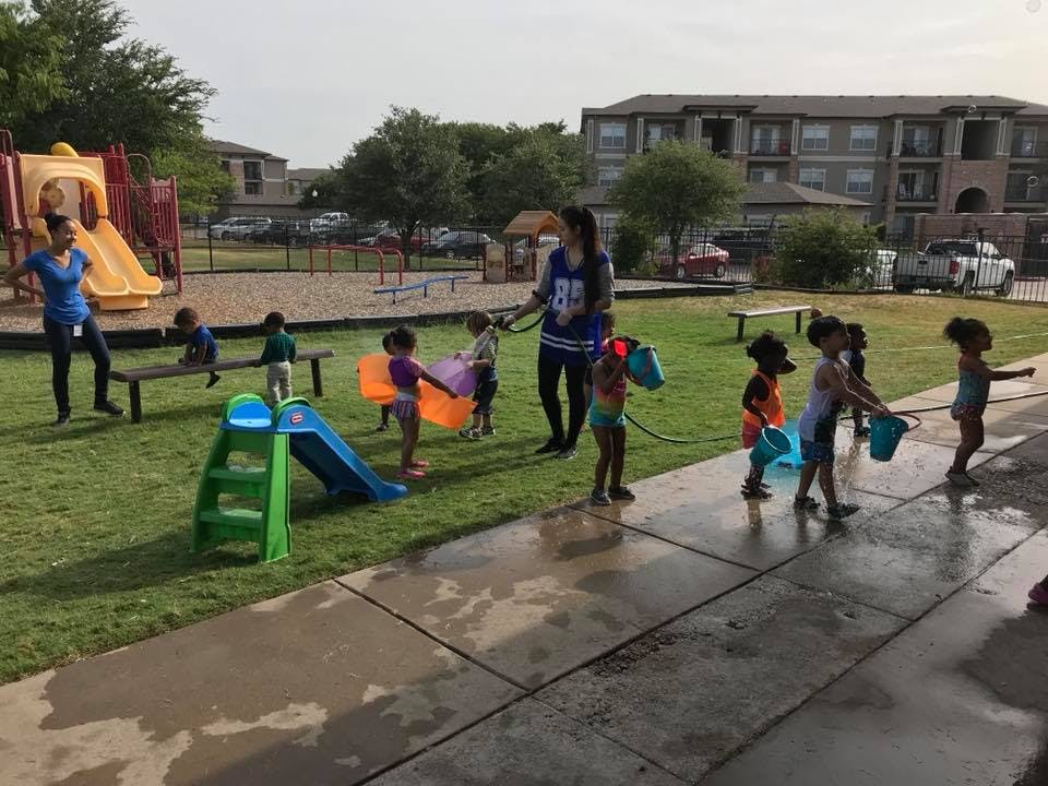 People and children playing with water in a grassy area with a playground