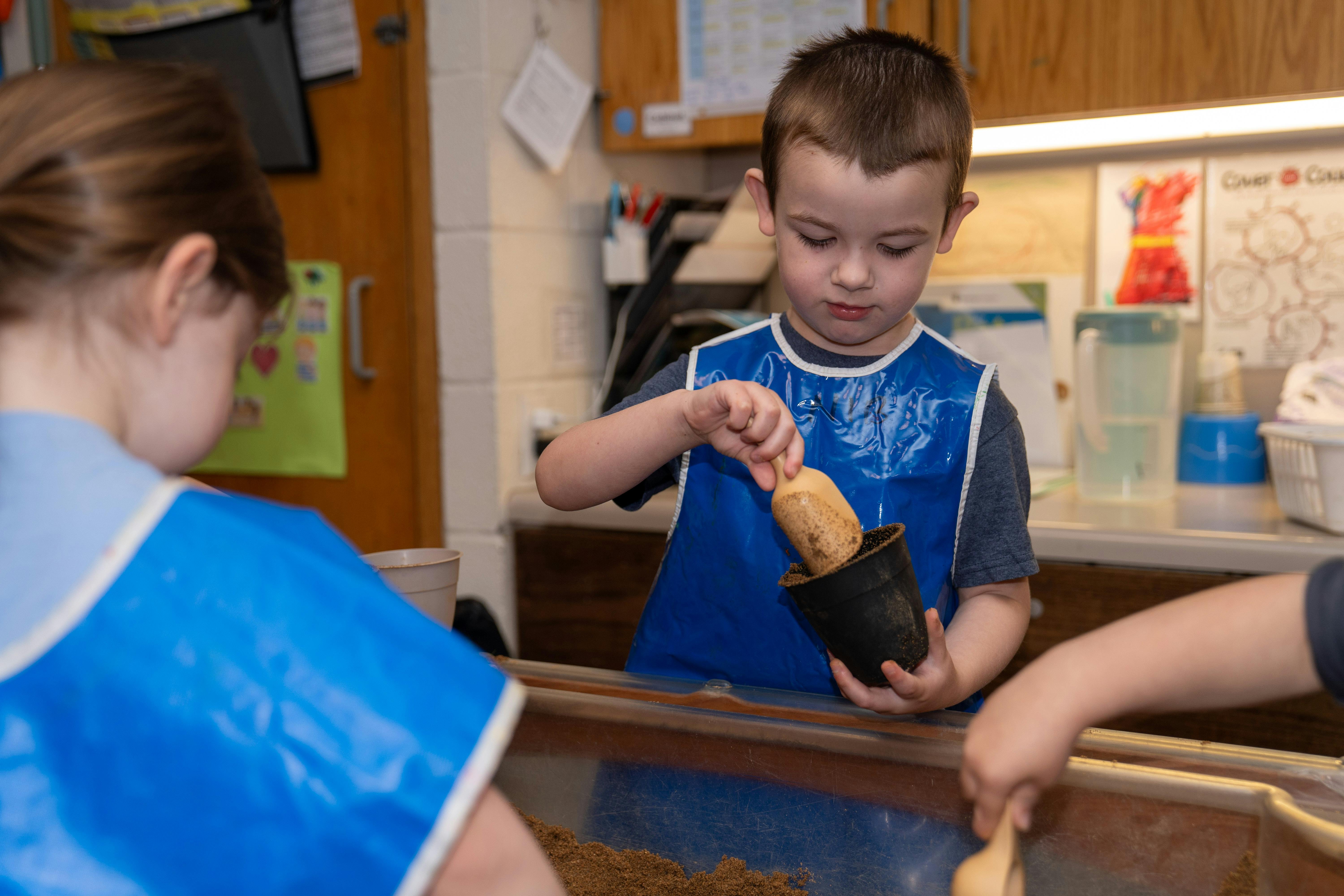 Child in blue smock scooping sand into a small black pot