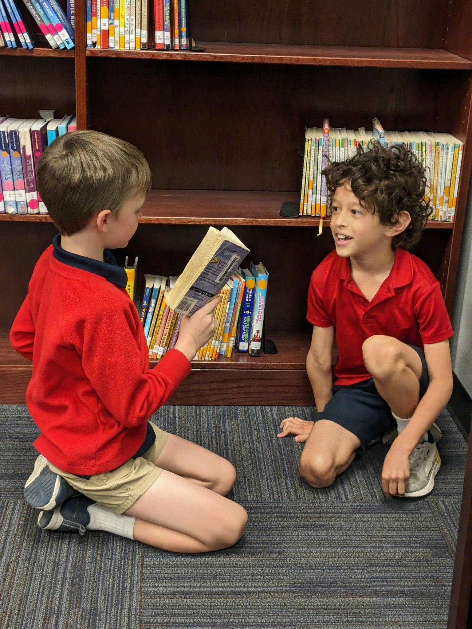 Two children kneeling on the floor in front of a bookshelf, one reading a book
