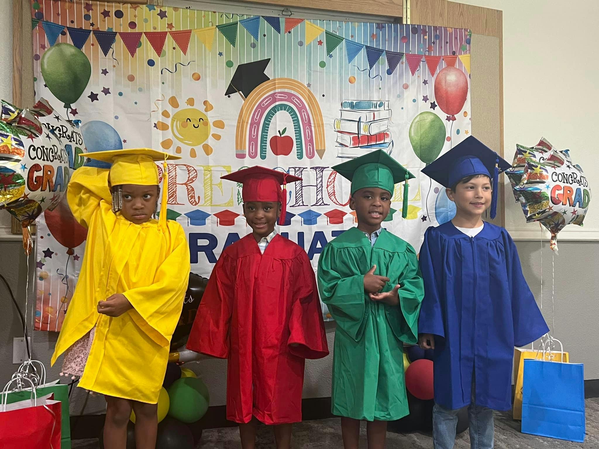 Four children in graduation caps and gowns standing in front of a colorful backdrop