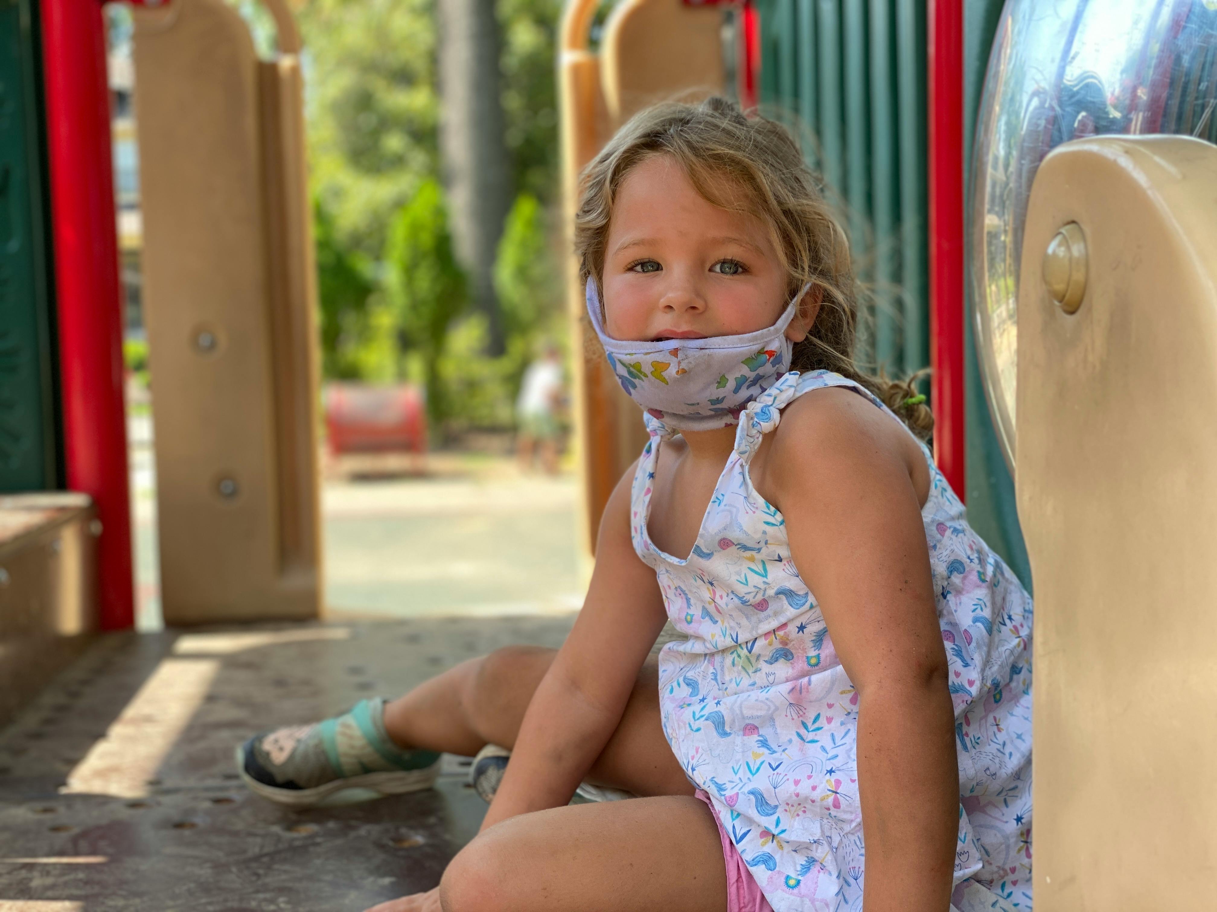 Child wearing a face mask pulled down, sitting on a playground
