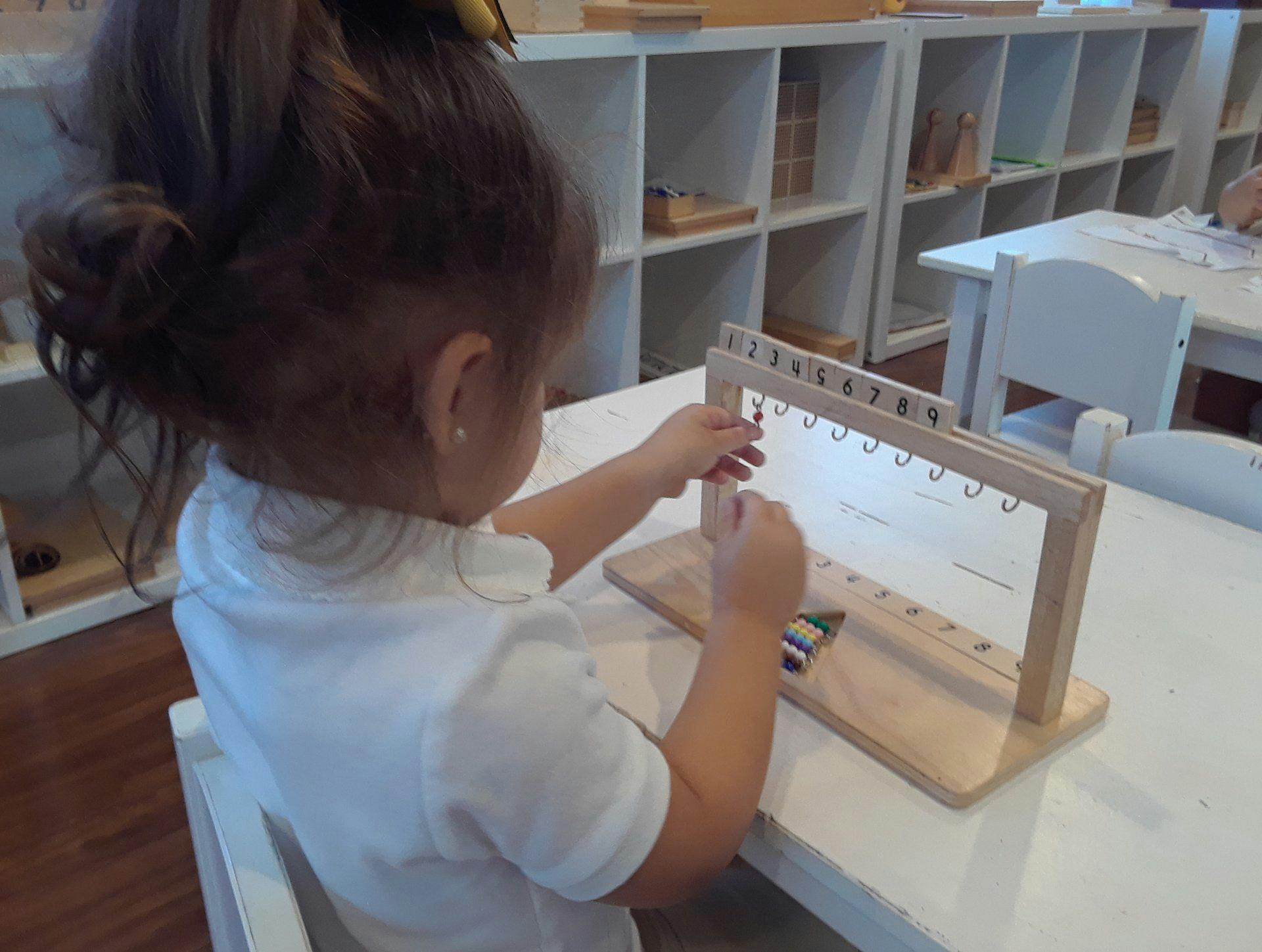 Child manipulating a wooden frame with hooks and rings insets on a table in a classroom
