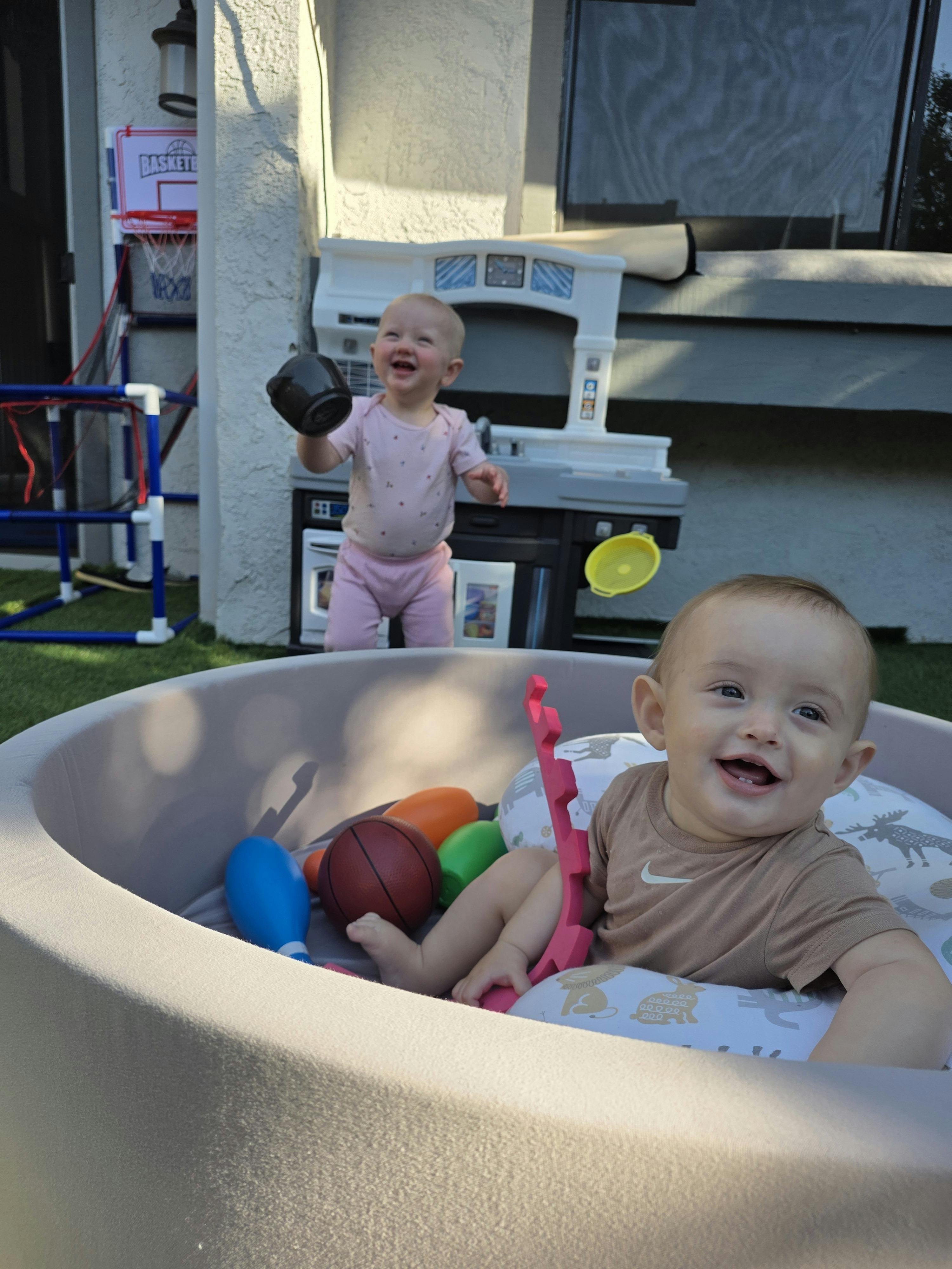 A child sitting in a soft playpen with another child standing behind them playing