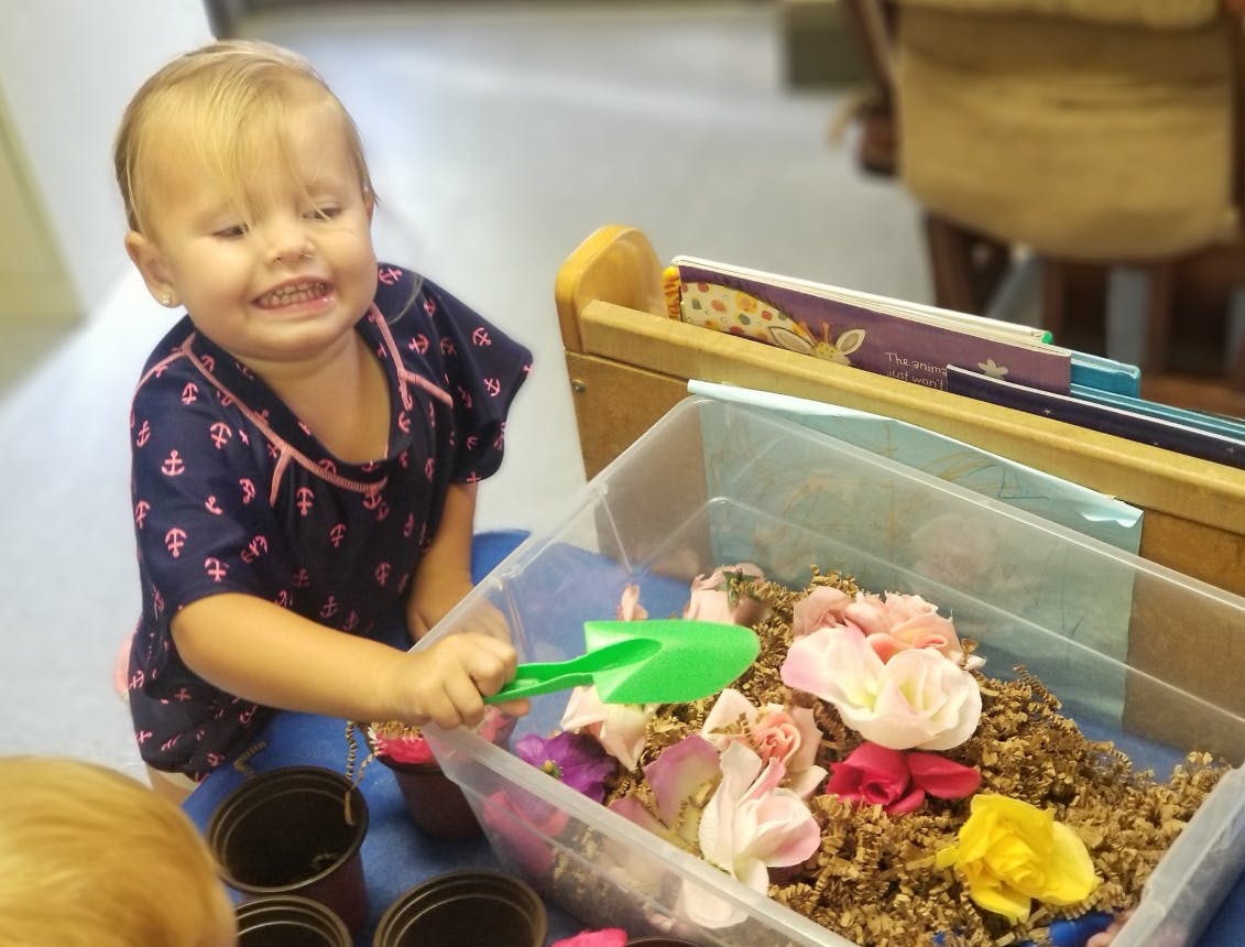 Child playing with a green shovel in a bin of dirt and flower petals