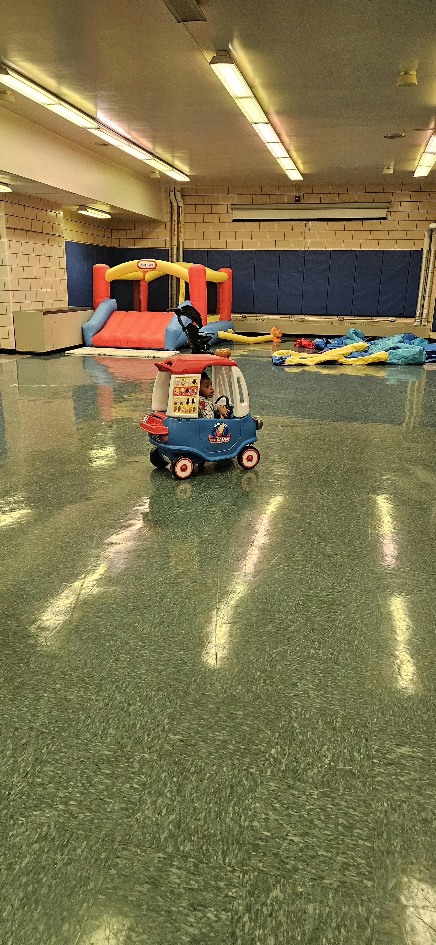 Child in a toy car in a room with an inflatable bounce house