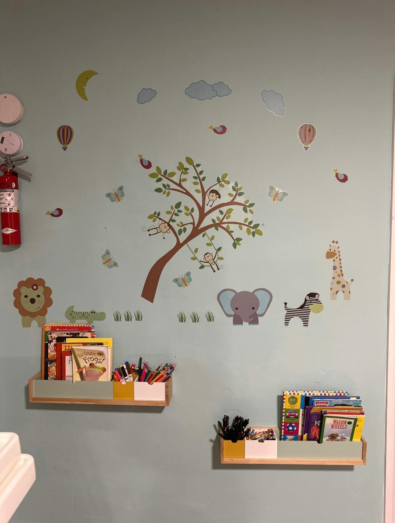 Wall decorated with animal and tree decals, with two shelves of books and art supplies