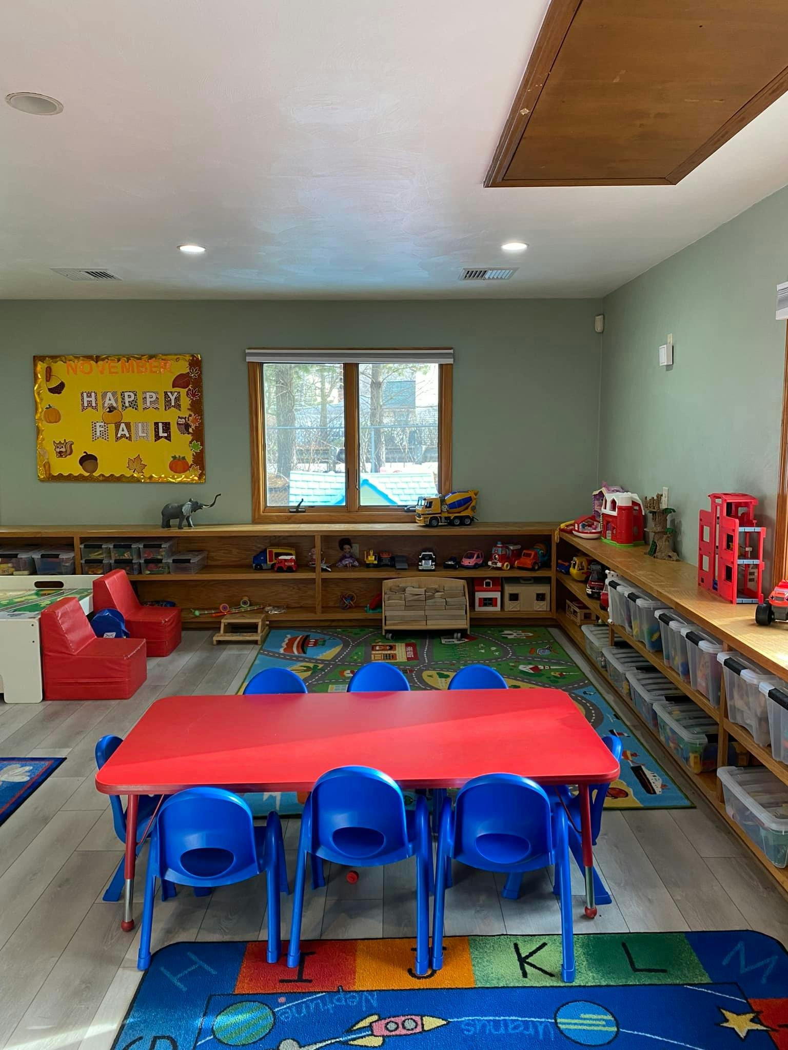 Classroom with a red table and blue chairs, shelves with toys, and a window