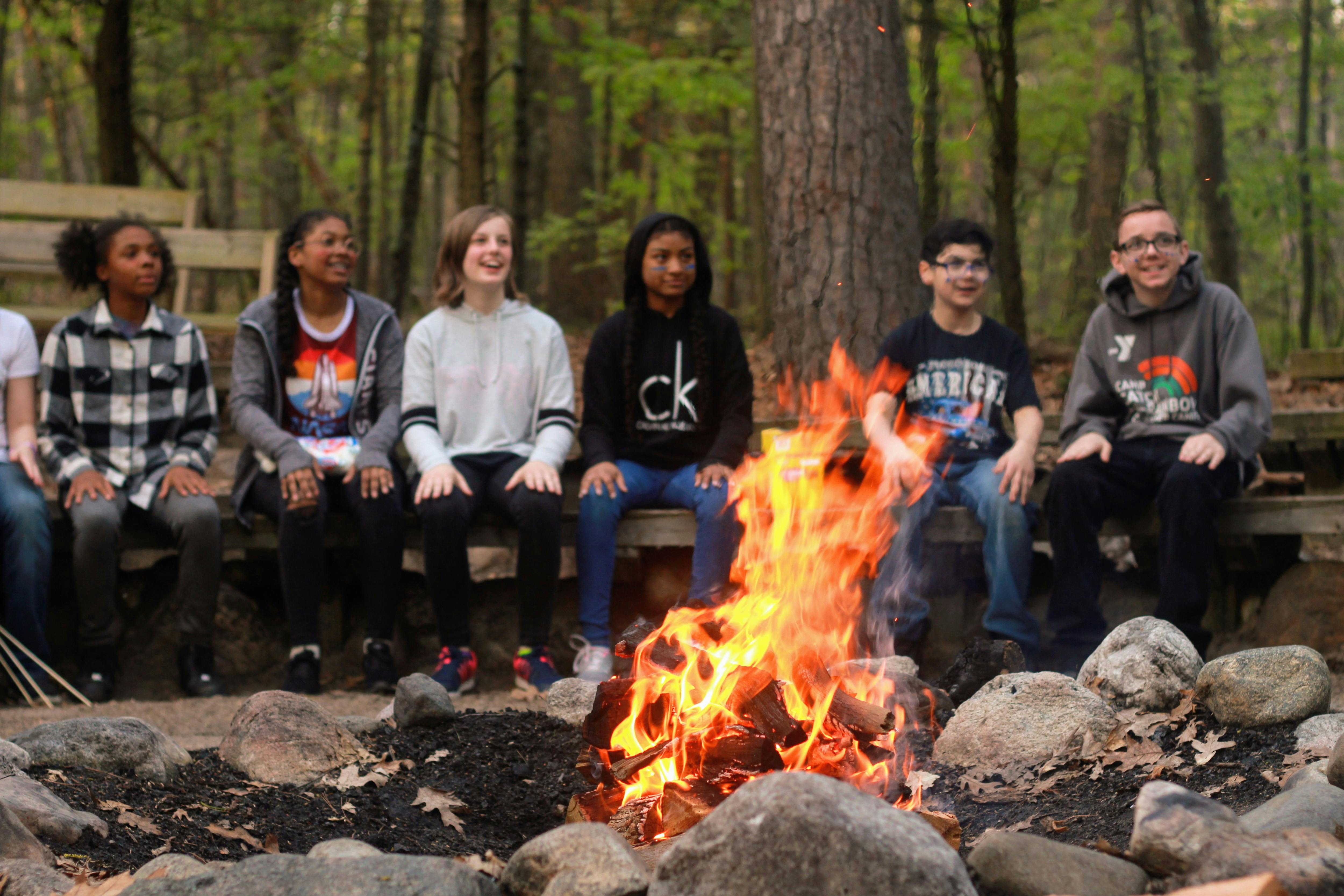 Six teens sitting around a campfire in a wooded area