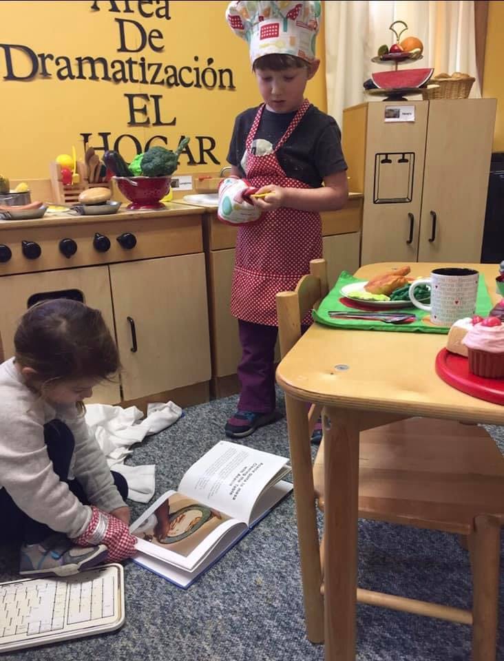 Child in chef hat and apron holding toy food, another child looking at a book