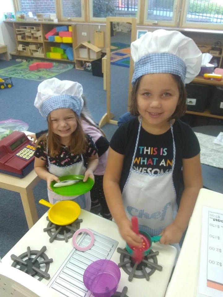 Two children wearing chef hats and aprons playing with toy kitchen