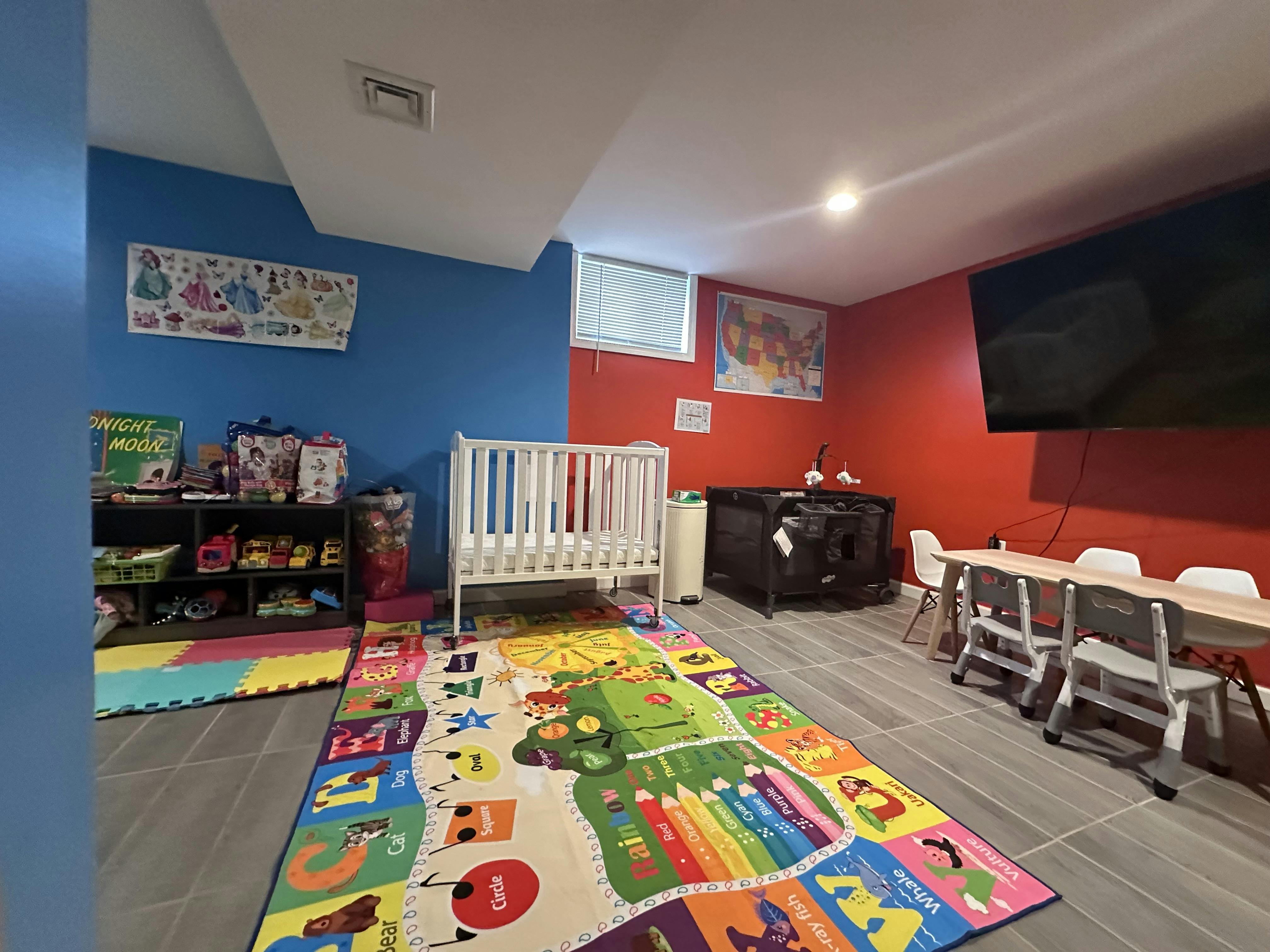 A childcare room with a crib, playpen, children's table and chairs, and a colorful alphabet rug