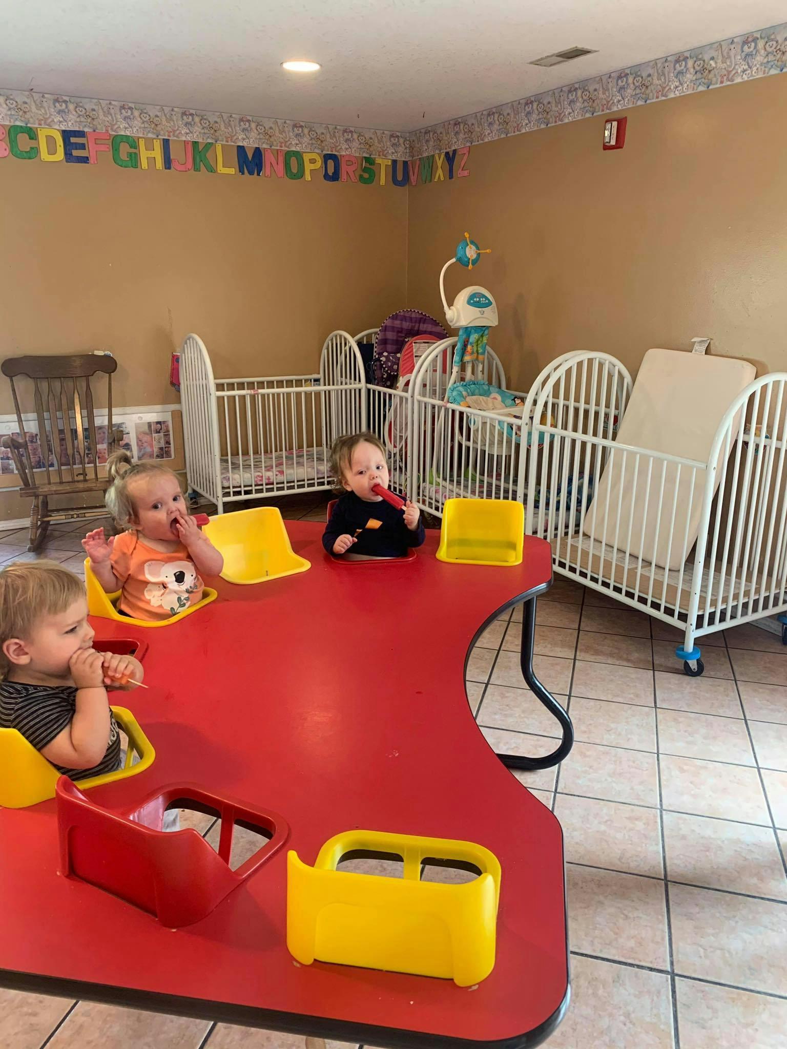 Three children sitting at a red table with cribs in the background
