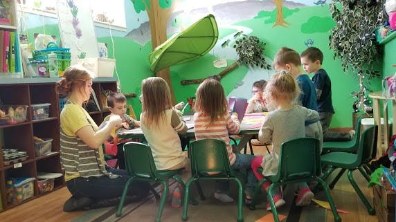 Person kneeling with children at a table in a classroom