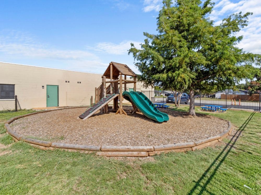 A playground with a wooden play structure and green slide on wood chips