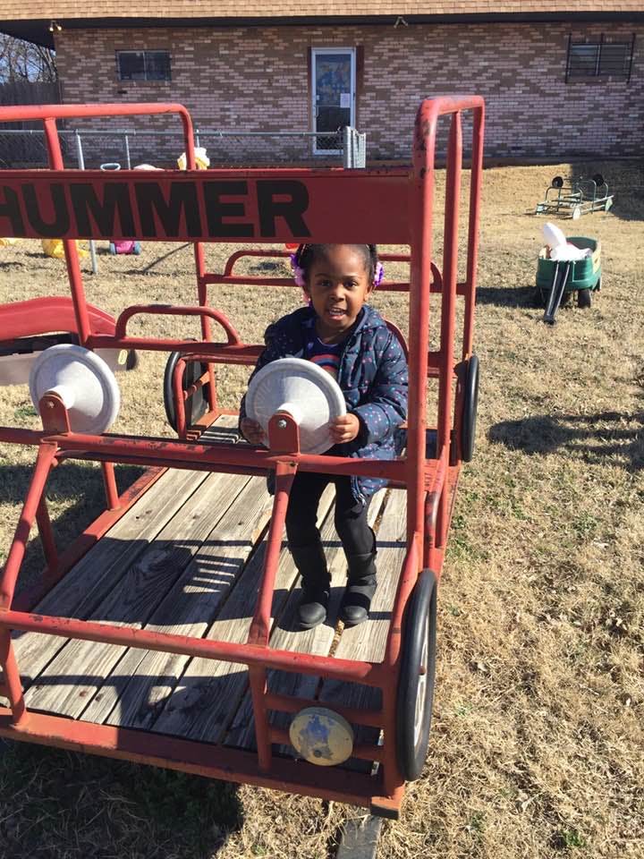 Child sitting in a red toy vehicle with "HUMMER" on the side