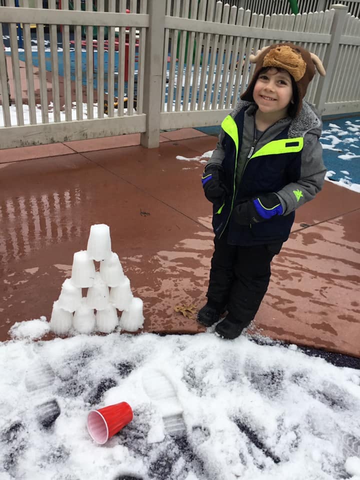 Child in winter clothing standing next to a pyramid of white cups in snow
