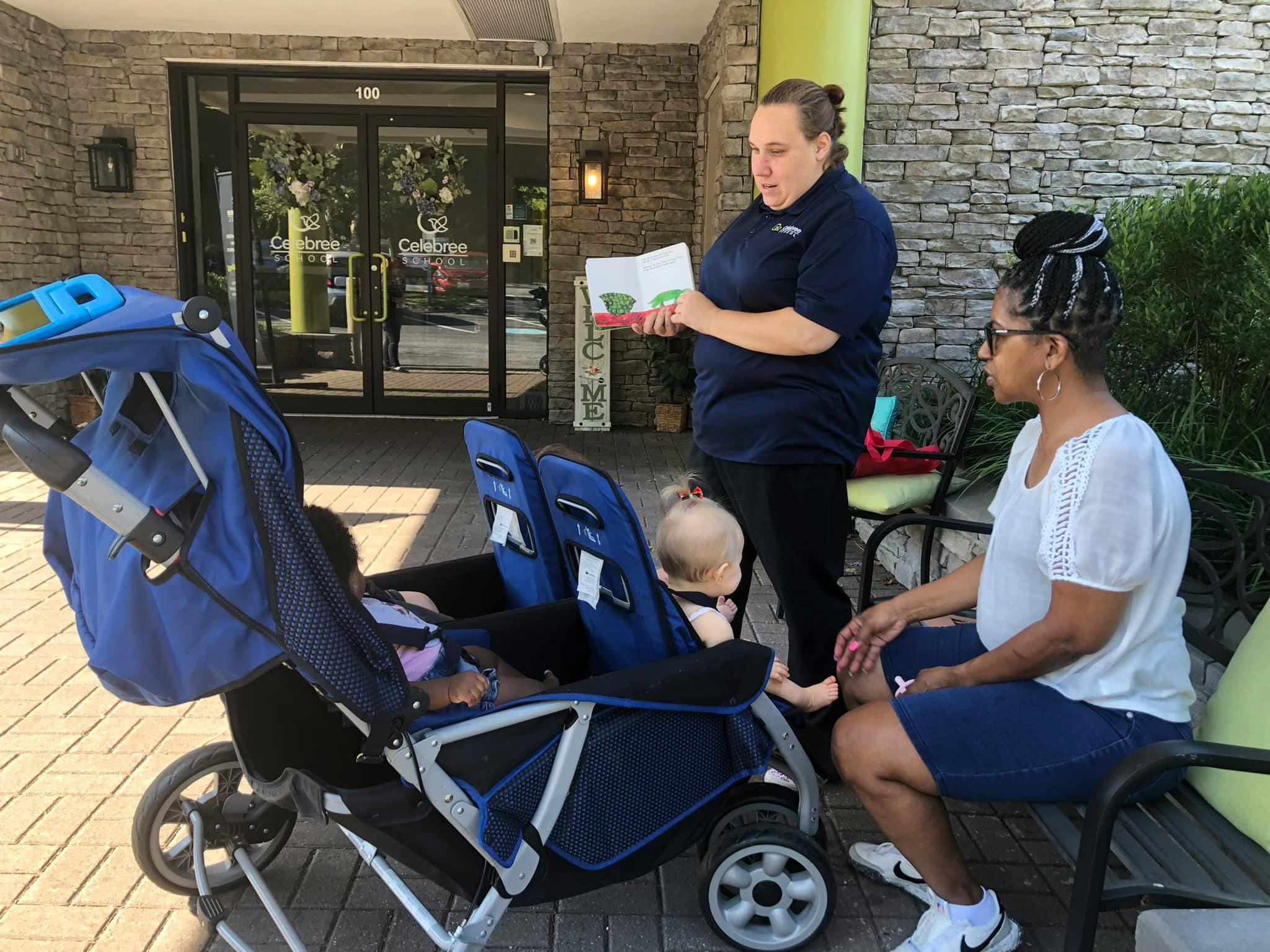 Person reading a book to a seated person and a child in a stroller