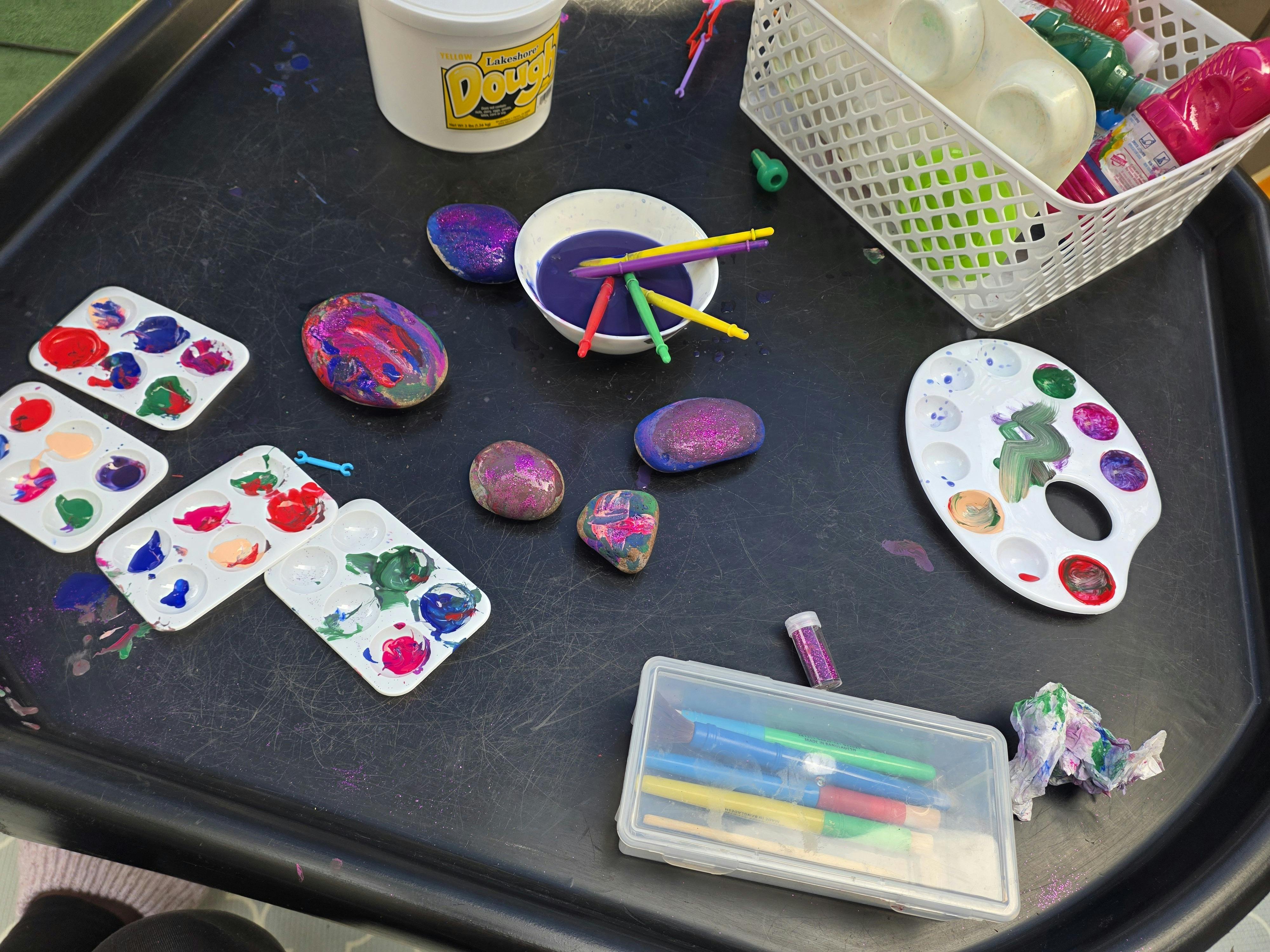 multiple paint palettes with various colors, painted rocks, brushes, and glitter