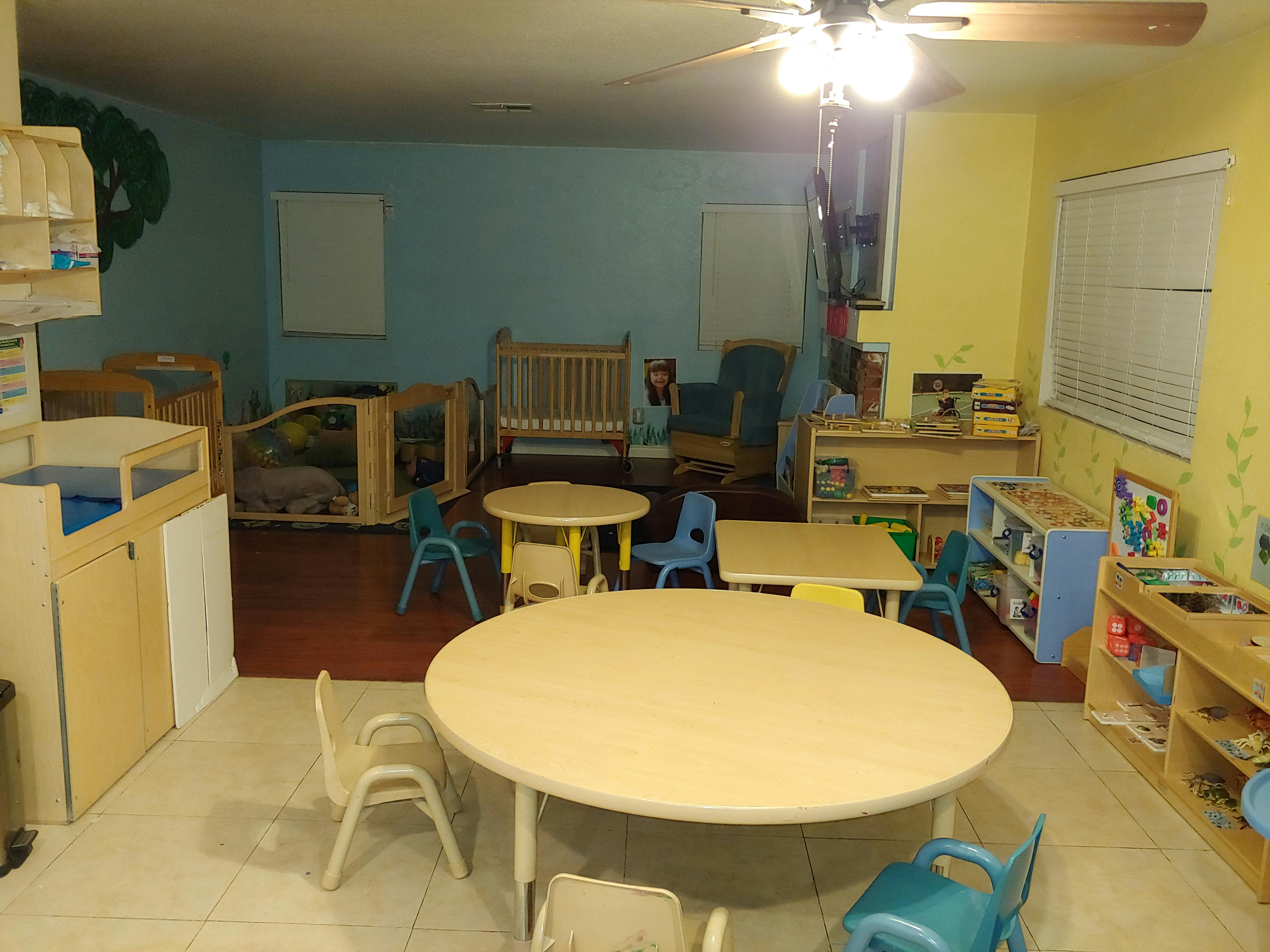 A classroom with small tables and chairs, cribs, and shelves with blue and yellow walls