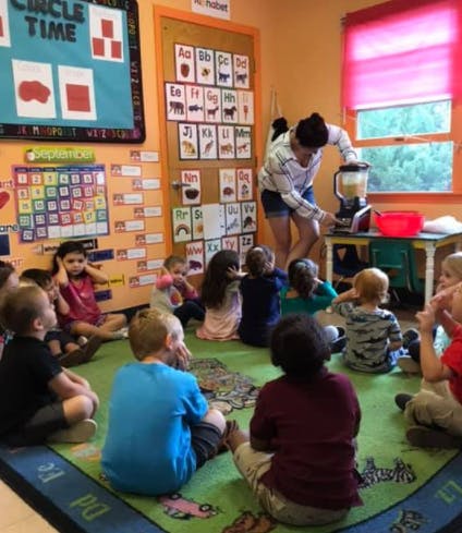 Person using a blender in a classroom with children sitting on the floor