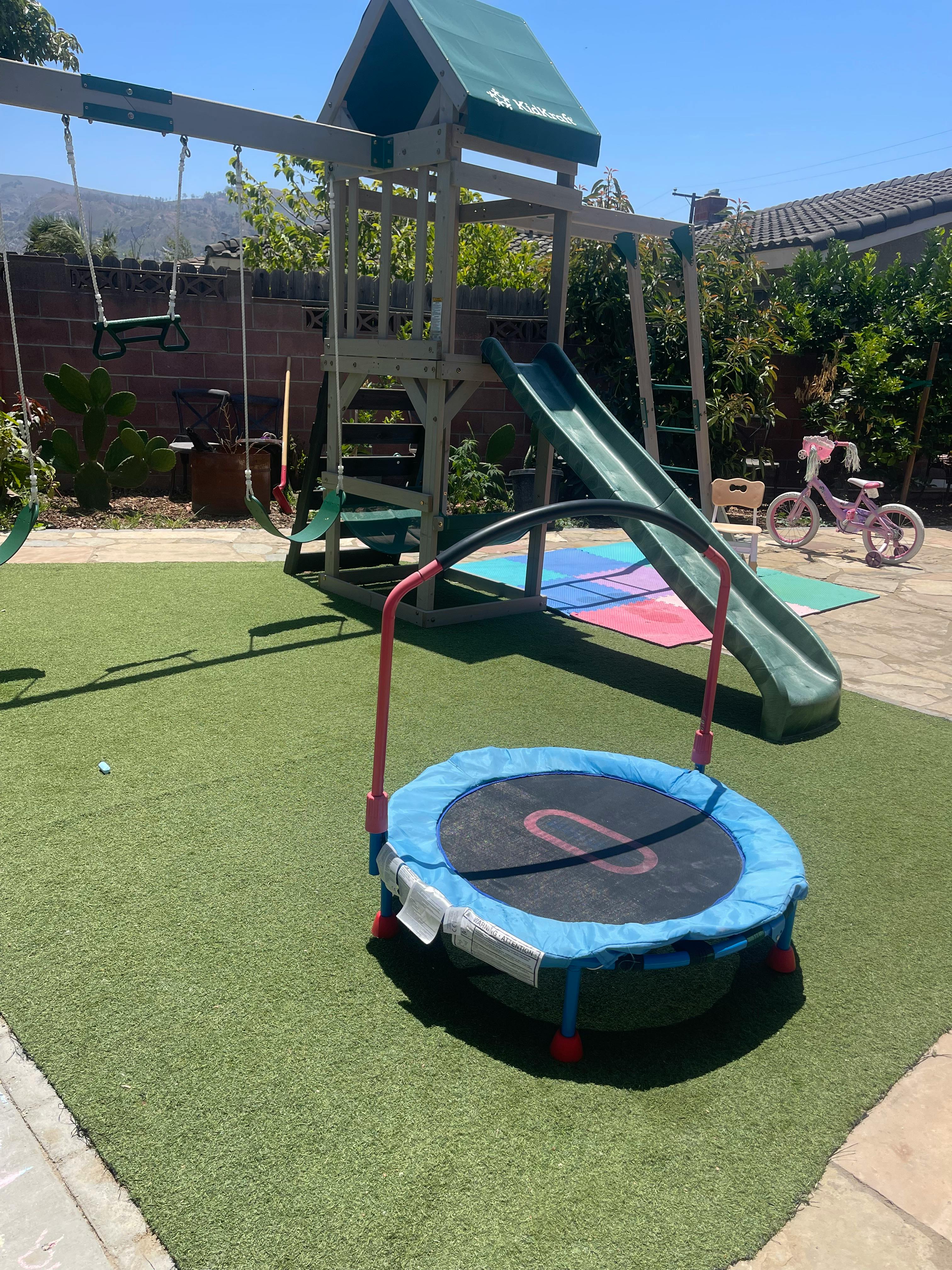 Playground with a slide, swings, and a small trampoline on artificial grass