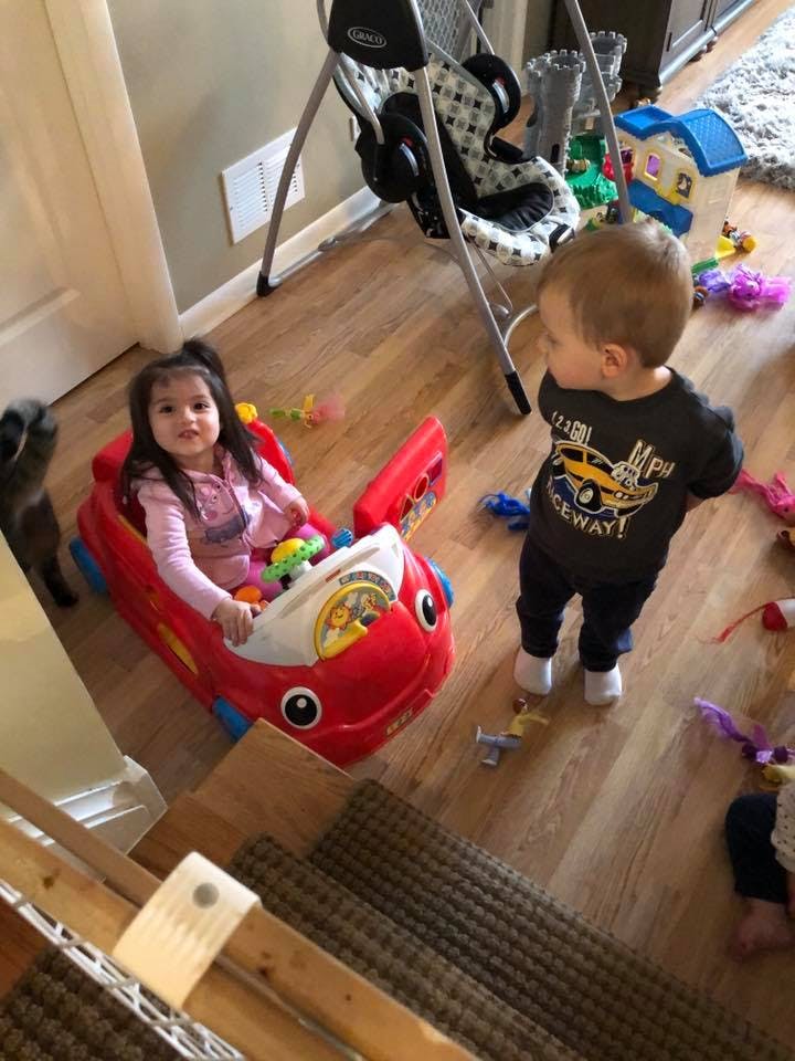 Child sitting in a toy car and a child standing nearby