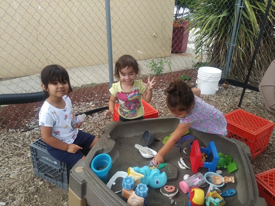 Three children playing with toys in a sand and water table outdoors