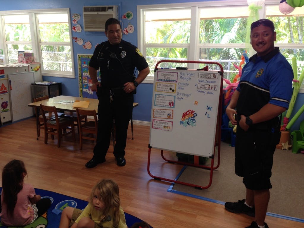 Two people in uniform standing in a classroom with children sitting on the floor