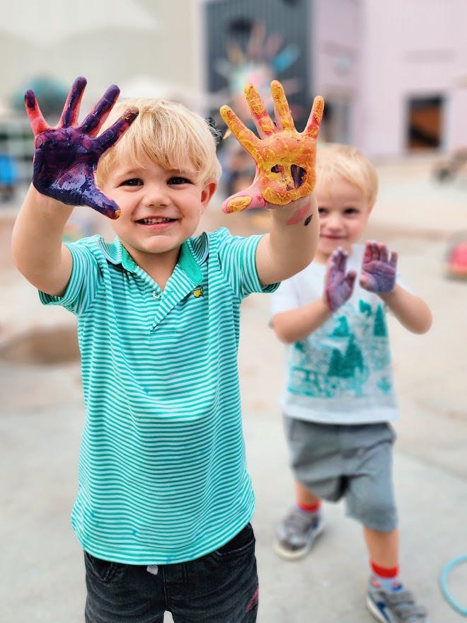 Child smiling with paint-covered hands held up, another child in background