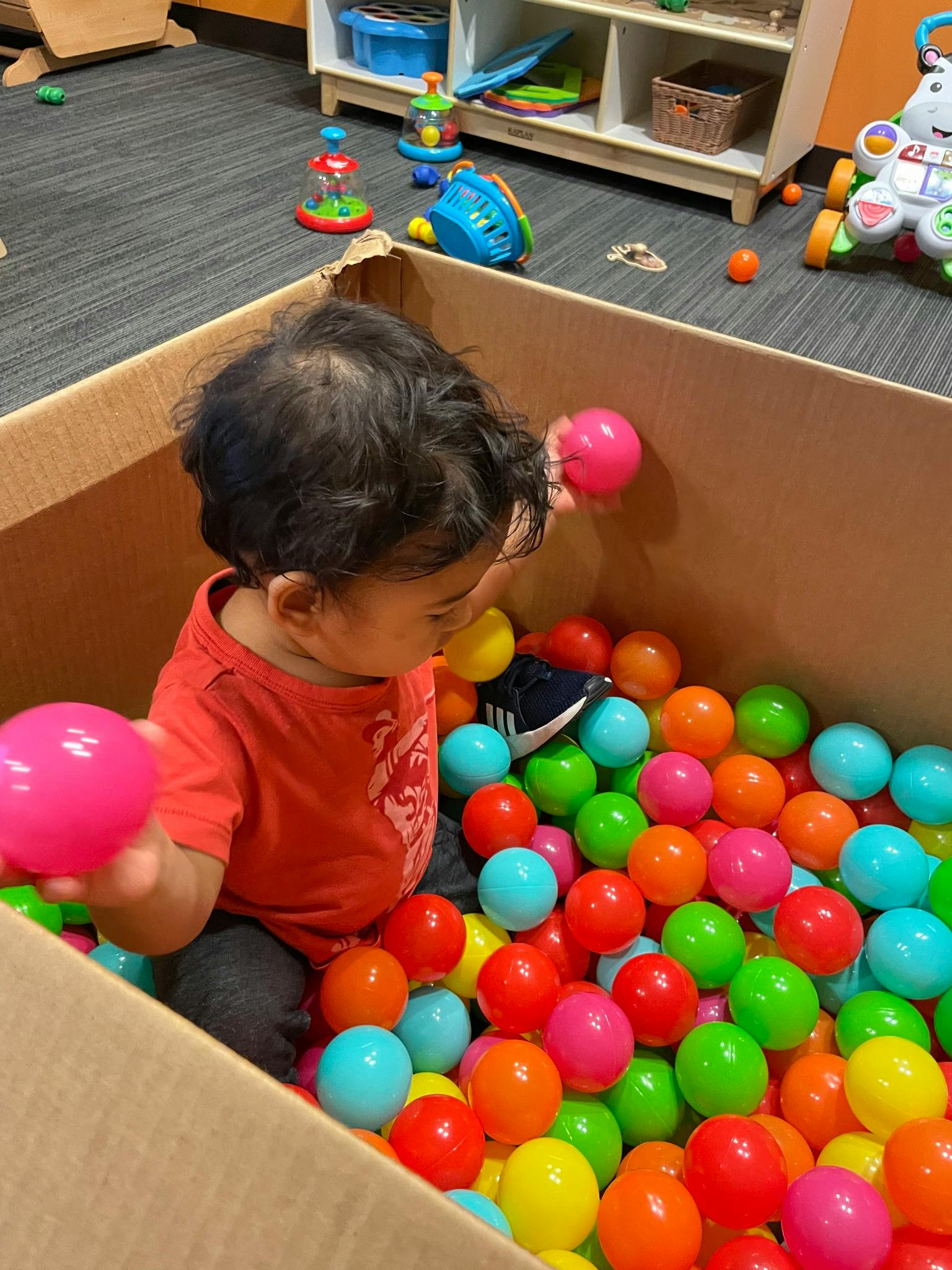 Child playing in a cardboard box filled with colorful plastic balls