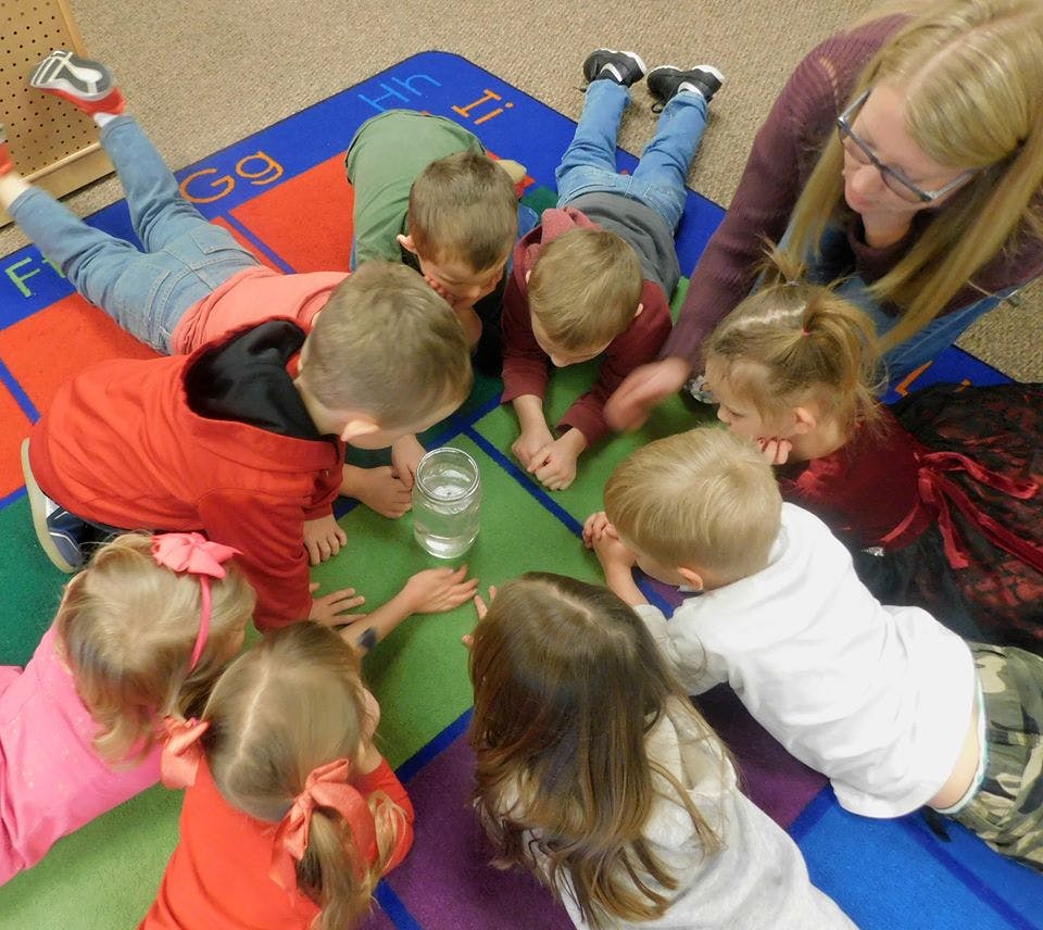 Adult and children gathered around a glass of water on a colorful rug