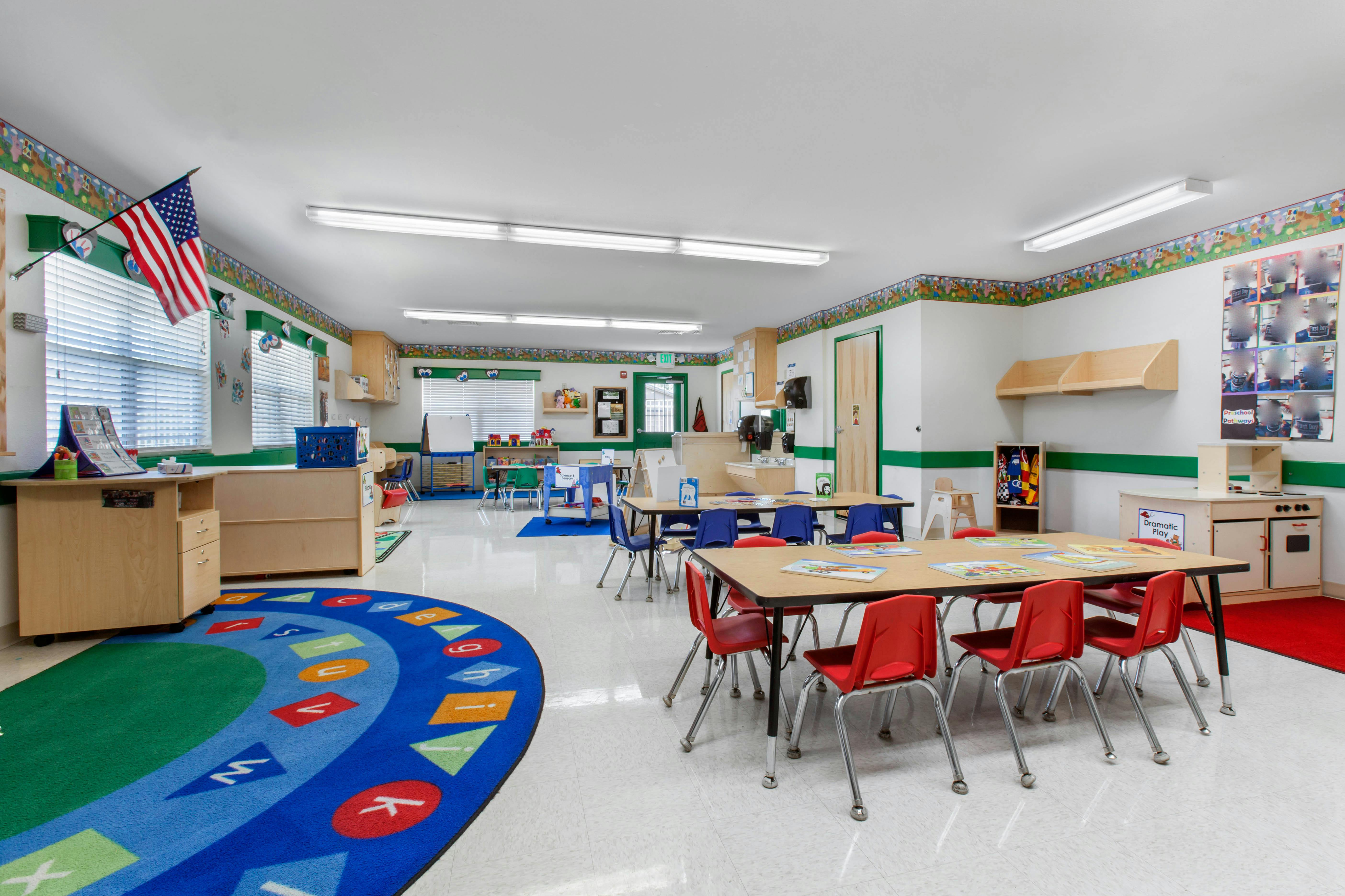 Classroom with child-sized tables and chairs, a play kitchen, and an alphabet rug