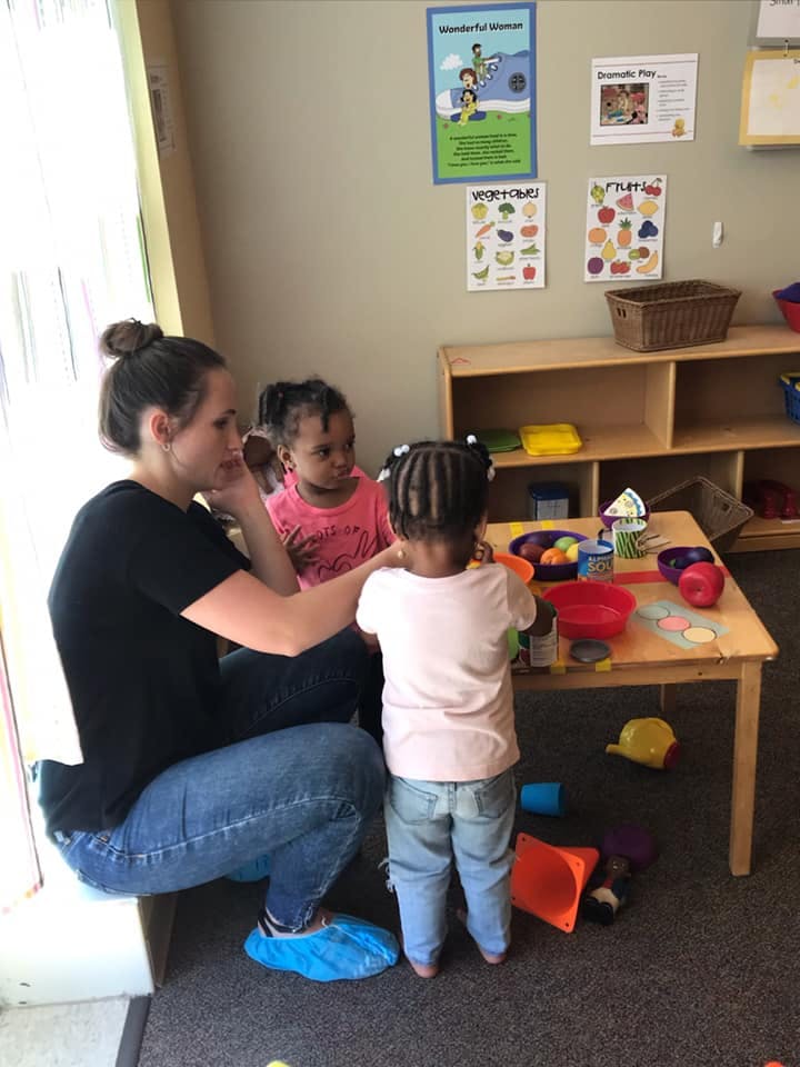Person crouching with two children playing at a small table in a classroom