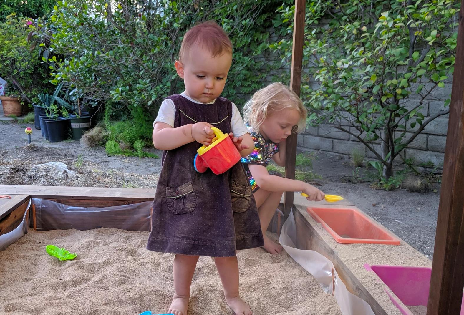 Two children playing in a sandbox, one holding a toy watering can
