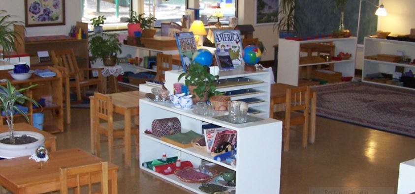 Classroom with small wooden tables and chairs, white shelves with learning materials
