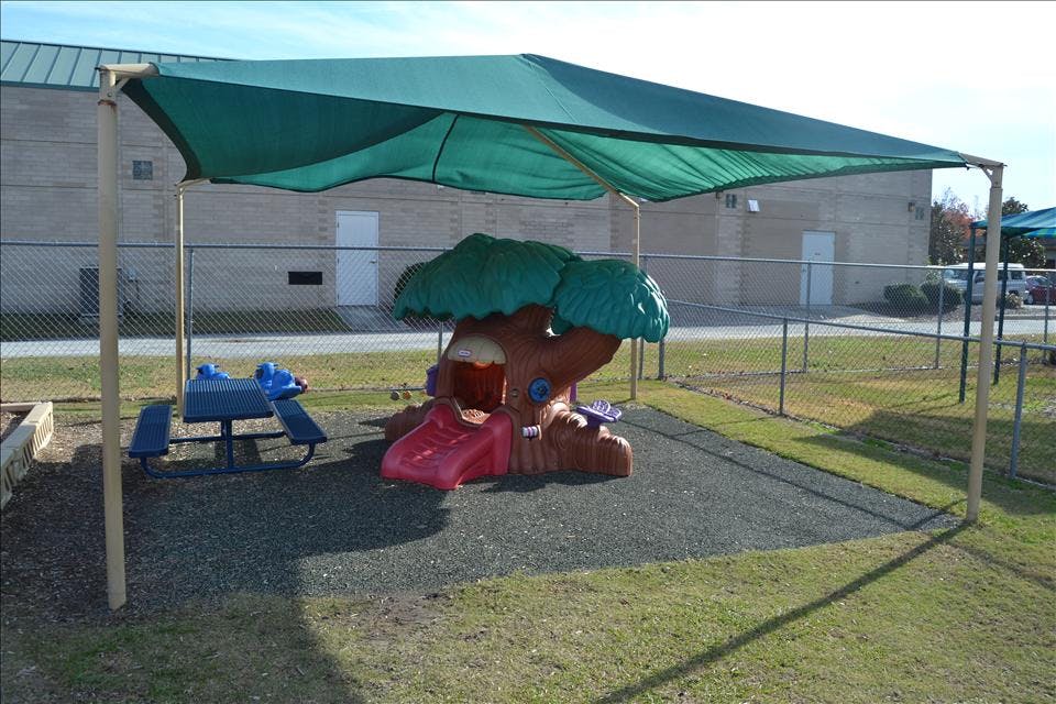 Playground area with a green shade cover, a tree-shaped playhouse with a slide, and a blue picnic table