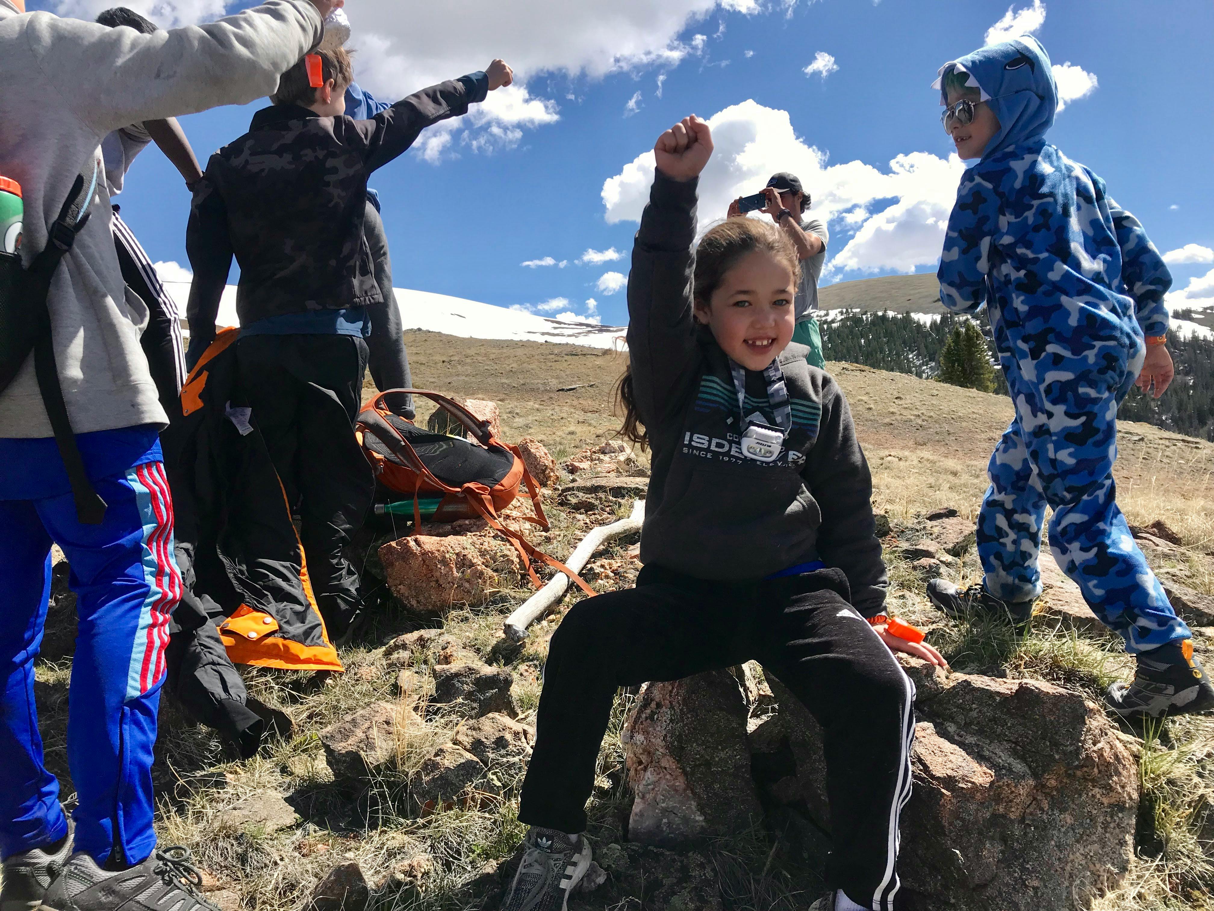 Child sitting on a rock with arm raised, other people standing around