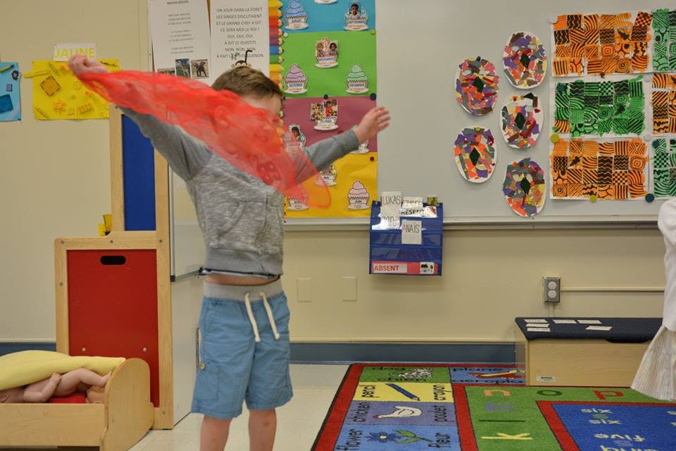 Child holding a red scarf with arms outstretched in a classroom